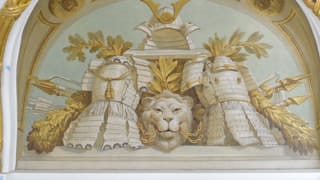 A photo of a painting near the ceiling in Asahi no Ma. A helmet and two suits of armor are depicted, and between them, a lion's head biting chains. Behind these are arrows, javelins, and lances.