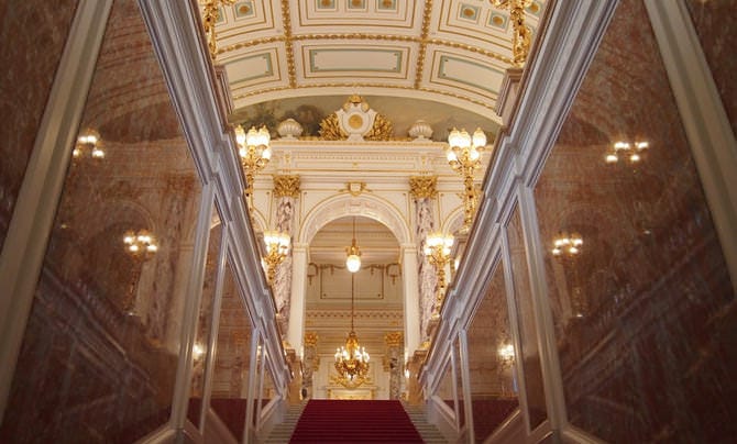 Una fotografía de la escalera central. Se ha extendido una alfombra roja por las escaleras, y las paredes a izquierda y derecha están revestidas con reluciente mármol rojo. Hacia delante, cerca del techo, se puede observar el emblema del crisantemo imperial.