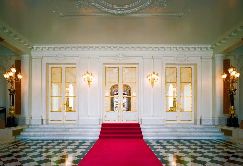 A photo of the entrance hall. The floor has a black and white checkerboard pattern, and a red carpet has been laid in its middle, extending towards the central staircase.