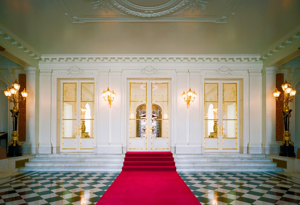 Una fotografía del vestíbulo de entrada. El suelo tiene un patrón de damero en blanco y negro, se ha extendido una alfombra roja en su centro, que continúa hasta la escalera central. 