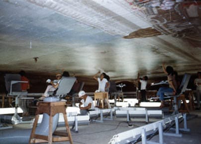 Una imagen de los trabajos de reforma que comenzaron en la Casa de Invitados del Estado, Palacio de Akasaka, en 1910. Para restaurar las pinturas en el techo de la sala Asahi no Ma, muchas personas están sentadas en sillas, mirando hacia el techo, trabajando sobre una plataforma construida sobre un andamio cerca del techo.