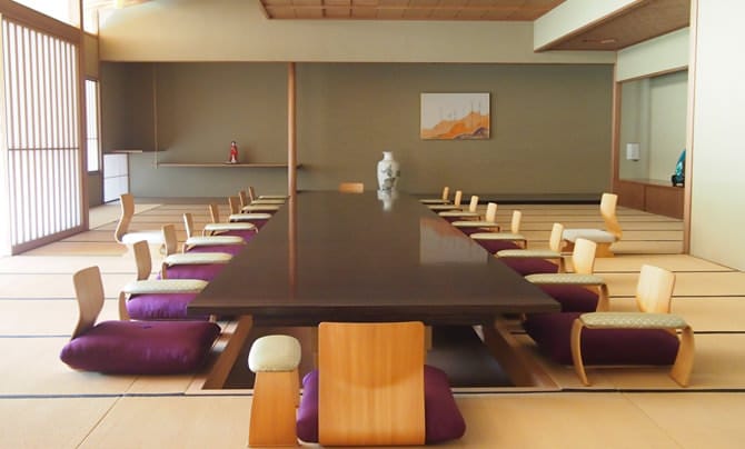 Une photo de la salle principale de style japonais de l’annexe japonaise. Une table a été placée au-dessus d’un espace en creux pour les pieds dans cette spacieuse salle de tatamis tandis qu’au fond, on peut voir un toko-no-ma (alcôve pour placer des fleurs ou une calligraphie) d’un noir de jais.