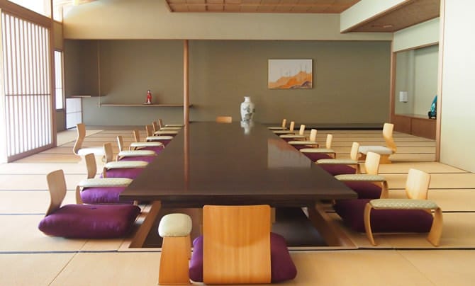 A photo of the main Japanese-style room in the Japanese Annex. A table has been placed over a sunken foot space in this spacious tatami room, and in the distance one can see a toko-no-ma (alcove for displaying flowers or calligraphy) in jet black.