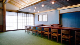 Una fotografía de las sillas de comedor del anexo japonés. Las sillas se han colocado junto a la barra. Las columnas y vigas expuestas de la sala están hechas con robusta madera de castaño, y se han colocado listones de bambú en el techo, creando una apariencia rústica.