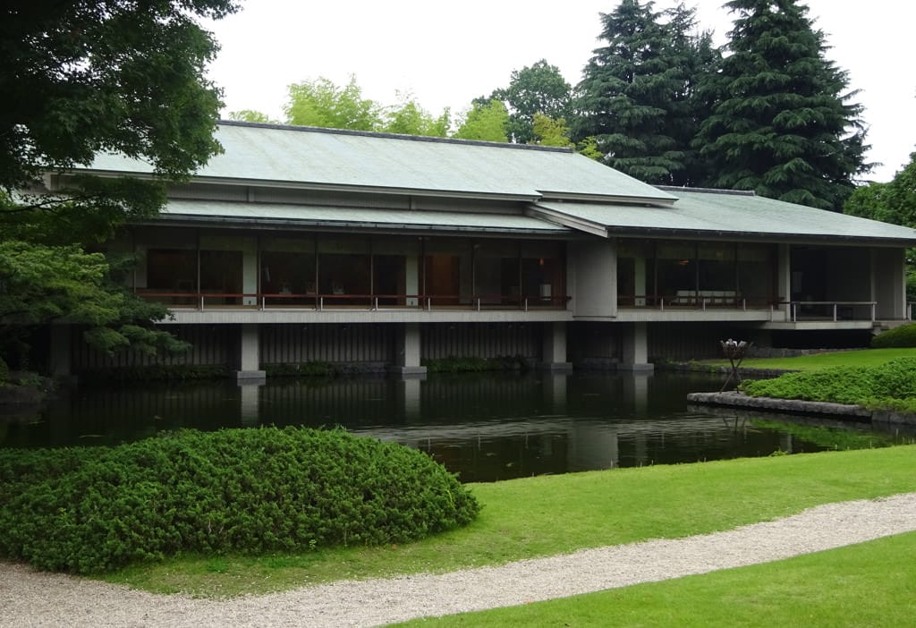 A photo of the exterior of Yushintei, the Japanese Annex. There is a path through the lawn leading to the annex. Across a pond, the gabled roof of the one-story Japanese Annex can be seen.