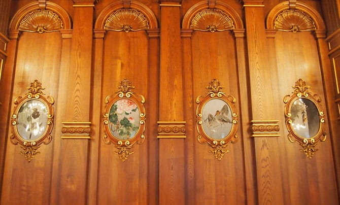 Une photo de l’œuvre en cloisonnés encastrés dans les murs de la salle Kacho no Ma. De gauche à droite, les panneaux cloisonnés représentent respectivement : « Pie et cornille », « Rougequeue aurore et pivoines », « Sarcelle d’hiver et roseaux » et « Pic rouge et cyprès ».