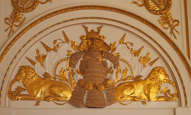 A photo of a gilded stucco relief of an armored warrior. The design is of the warrior is at center, with seated lions facing outwards on both sides.