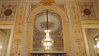 A photo of the great mirror in Sairan no Ma. Gilded stucco reliefs ornament the area around the mirror. The chandelier and ceiling decorations can be seen reflected in the great mirror.