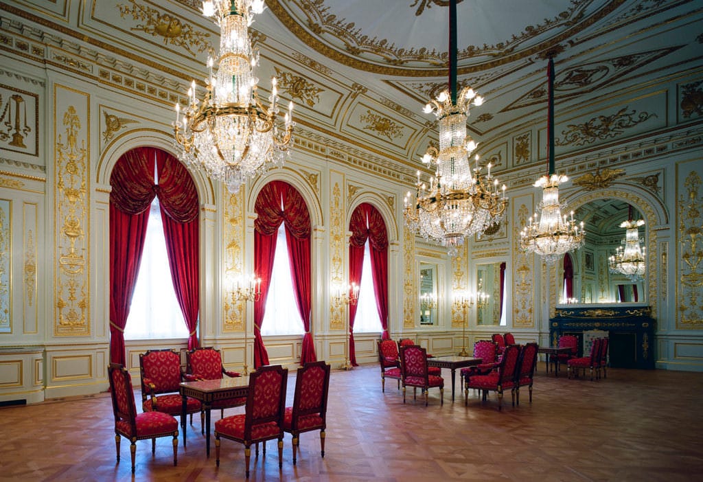 Una fotografía de vista completa de la sala Sairan no Ma. Tres lámparas de araña, espléndidos relieves de yeso recubiertos con pan de oro, y sillas y mesas ornamentales están presentes en la sala. Se tejen motivos en la tela roja de las sillas.