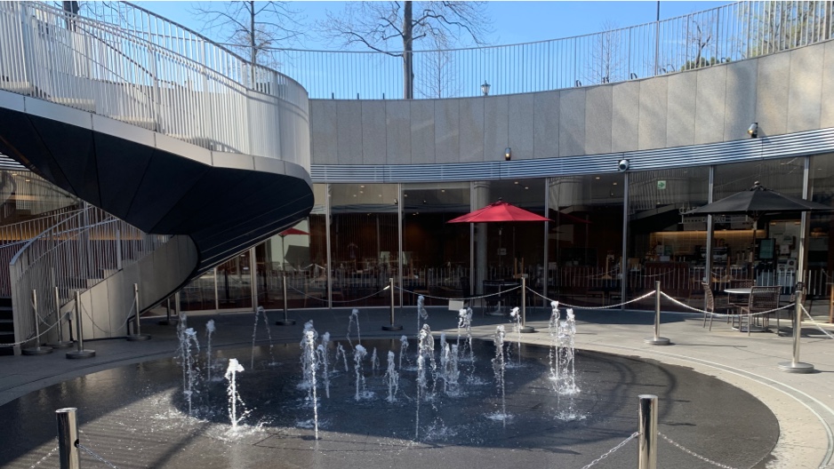 A photo of a central fountain ringed by the windows of the rest area and shops. The fountain area is open to a blue sky, and a curved staircase leads back to ground level.