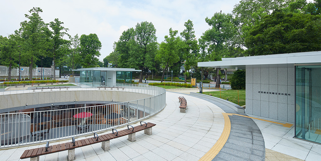 Foto del exterior del área de descanso de delante de la Casa de Invitados del Estado, Palacio de Akasaka Los árboles rodean una plaza central con bancos, y una gran abertura circular en el suelo desvela instalaciones subterráneas.