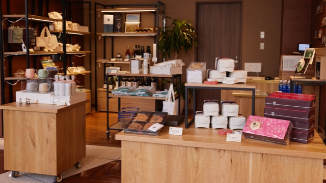 Foto del interior de la tienda. Tazas, dulces, botellas de vino y otros artículos se exponen en estanterías en un espacio sutilmente iluminado.