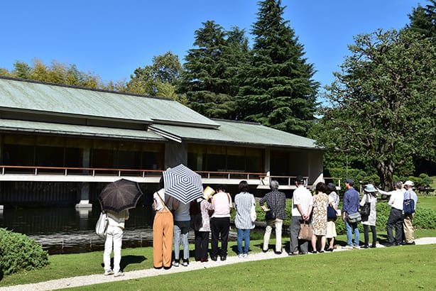 Une photo de visiteurs à l’annexe de style japonais, Yushintei. Les visiteurs sont alignés devant l’étang en face de l’annexe de style japonais, écoutant les explications de leur guide touristique. 