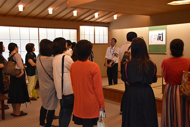 Una fotografía de los visitantes en la sala de té del anexo de estilo japonés, Yushintei. Los visitantes están escuchando la explicación del guía del recorrido durante su visita.