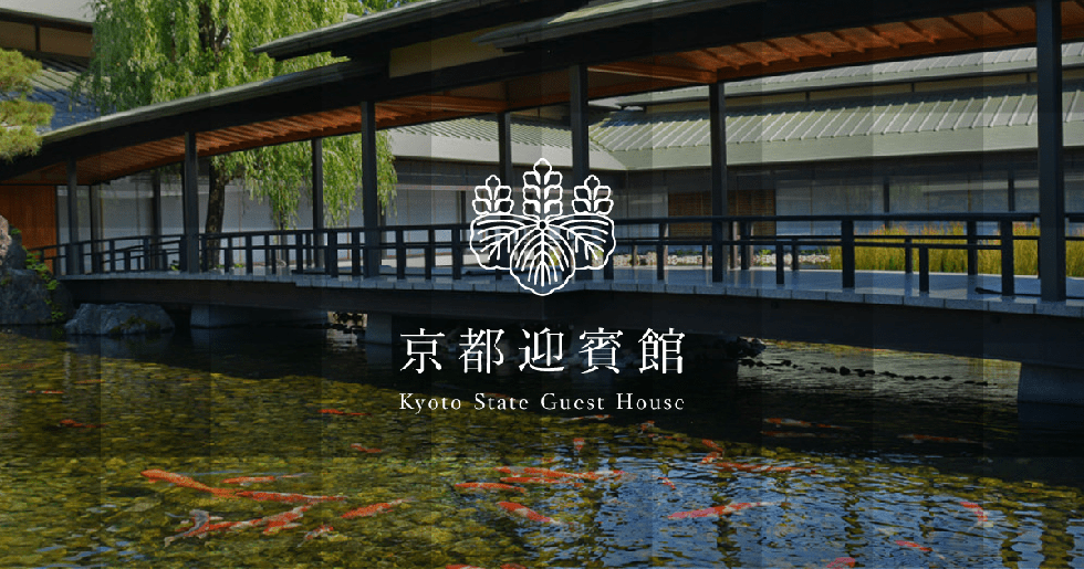 Dans le jardin et plus loin le long du bâtiment, des arbres verdoyants poussent dru sur un fond de ciel bleu sans nuages. Le ciel bleu se reflète dans l’étang calme au premier plan où de nombreuses carpes colorées nagent. Un large pont enjambe l’étang, reliant les crêtes est et ouest du toit de la Maison des hôtes d’État de Kyoto.