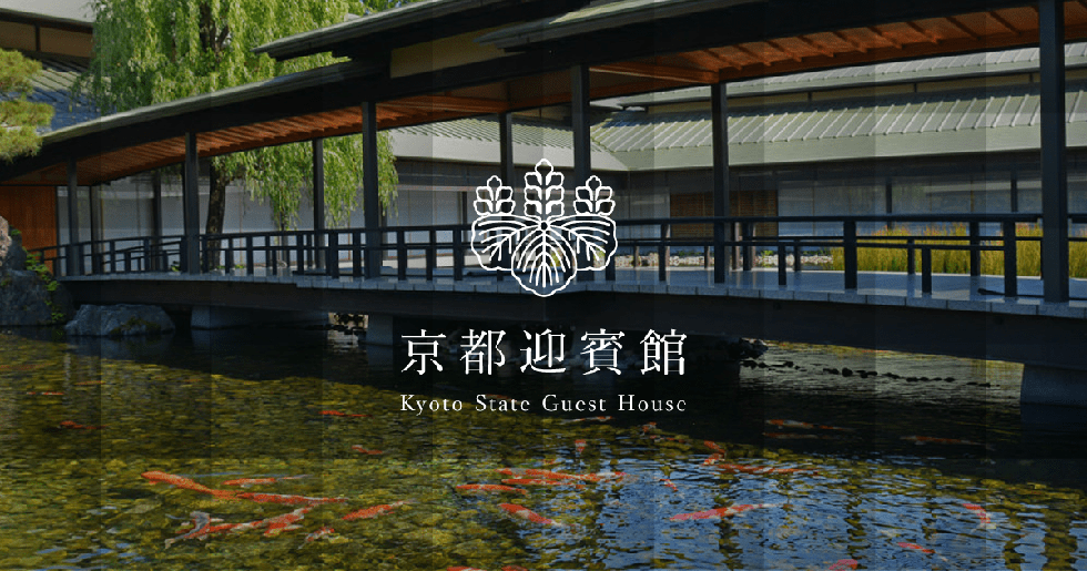 In the garden and further along the building, verdant trees grow thickly before a background of cloudless blue skies. The blue skies are reflected in the calm pond in the foreground, in which many colored carp are swimming. A broad bridge extends across the pond, connecting the eastern and western ridges of the roof of the Kyoto State Guest House.