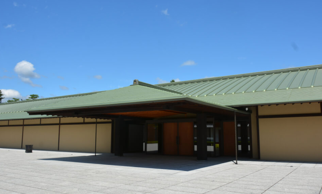 The front entrance is photographed here diagonally from the edge of the forecourt. The sky extends large and blue, and one can tell from this photo that the forecourt and the building are very large.