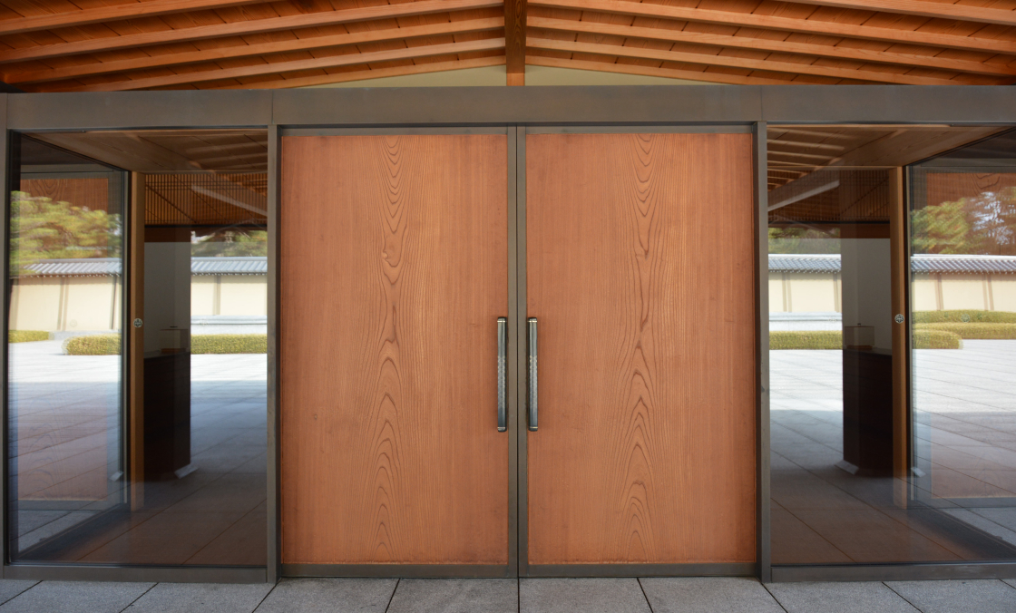 Les portes de l’entrée principale apparaissent au centre de la photo. Des vitres ont été installées de part et d’autre des portes, et le parvis s’y reflète comme dans un miroir.