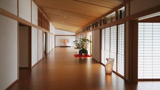 A corridor extends straight ahead. A kimono is on display at the end of the corridor, and in the middle of the corridor, before the front entrance is a flower arrangement. The flooring reflects the light from paper lanterns, and the light passing through the sliding paper doors of the hallway.