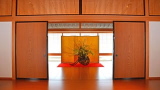 En esta fotografía, la puerta que conecta la entrada delantera con el corredor está abierta. Se ha colocado un arreglo floral tras las puertas. Un biombo dorado colocado detrás del arreglo floral, y una alfombra roja extendida debajo del florero. 