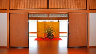 In this photo, the door connecting the front entrance and the corridor is open. A flower arrangement has been placed beyond the doors. A gold folding screen stands behind the flower arrangement, and a red carpet is spread below the flower vase. 