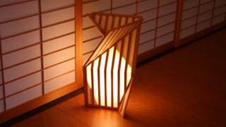 In this photo, a paper lantern set on the floor emits warm light in the dim hallway. Just behind the lantern are sliding paper doors. The lantern's design is evocative of origami.