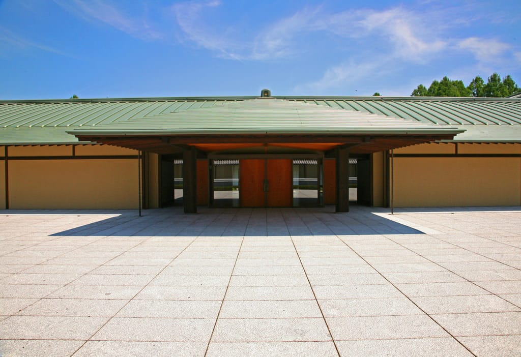 The front entrance appears with a background of blue skies. The green roof reflects the sunlight well. In the foreground, white stone tiles spread out in the forecourt.