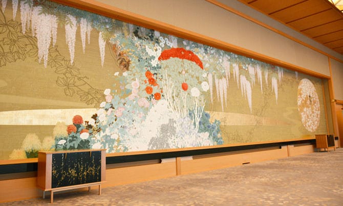 Une photo de la tapisserie « Reika » prise en biais. Comparé au présentoir placé devant, on peut constater la taille imposante de la tapisserie. Les plantes à fleurs de la tapisserie sont tissées dans des couleurs si vives qu'on pourrait les confondre avec celles d’un tableau.