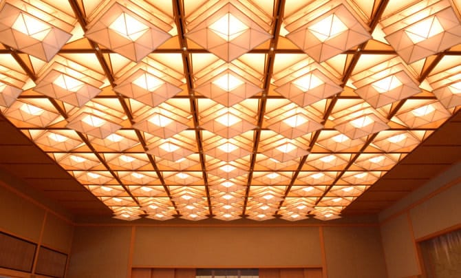 A photo of the illuminated coffered ceiling in Fuji no Ma. Rhombus-shaped lights have been installed in each delineated section of the ceiling, in shapes reminiscient of three-layered Japanese kites. Some lights extend out from the ceiling, facing downwards, and some do not extend, creating a pattern across the ceiling.