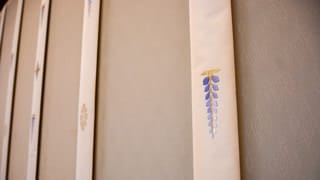 Una fotografía de un panel. Se han bordado flor de glicina, crisantemos, y lotos en las franjas de tela distanciadas de forma uniforme denominadas 
