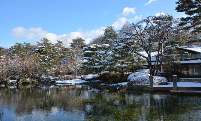 On voit le jardin en hiver. La neige s'est accumulée sur les arbres et le toit du bâtiment, dont l'image se reflète dans l'étang au premier plan.