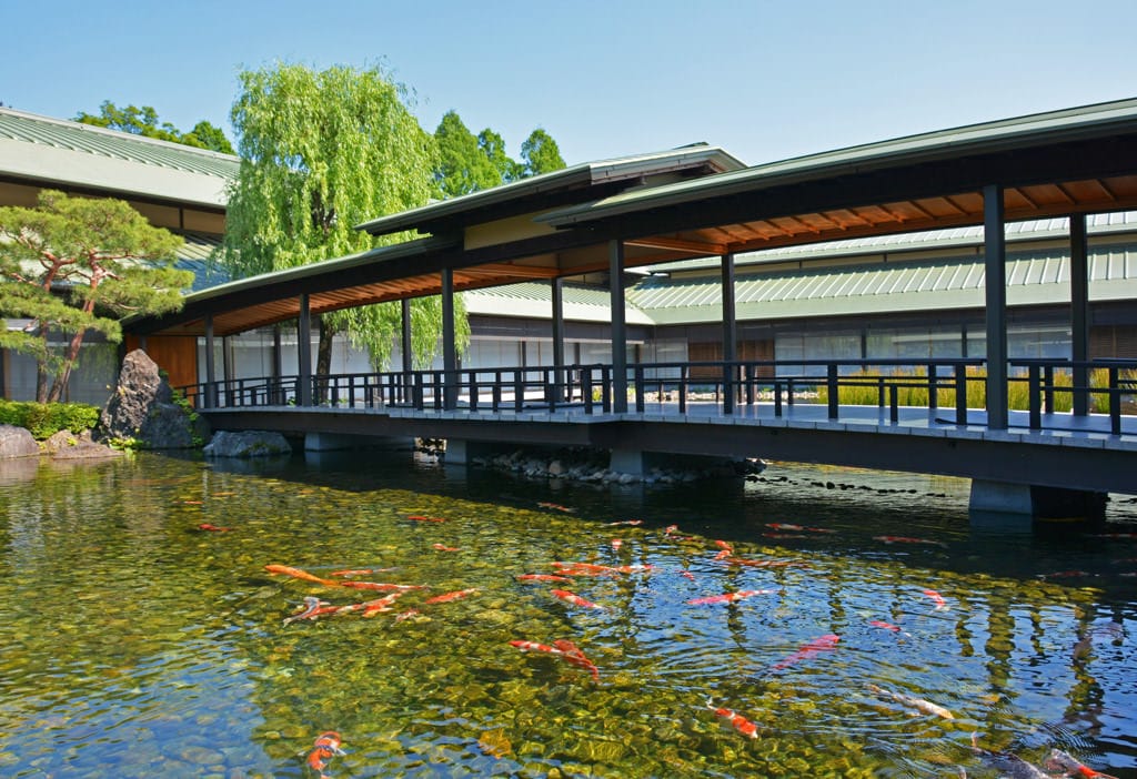 In the garden and further along the building, verdant trees grow thickly before a background of cloudless blue skies. The blue skies are reflected in the calm pond in the foreground, in which many colored carp are swimming. A broad bridge extends across the pond, connecting the eastern and western ridges of the roof of the Kyoto State Guest House.