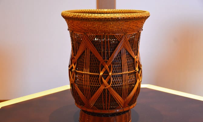 A woven flower vase is in the center of the photo. The vase's design is made with countless bamboo strips woven together. The vase is in the shape of a jar, but the top is made to open wide to accomodate flowers.