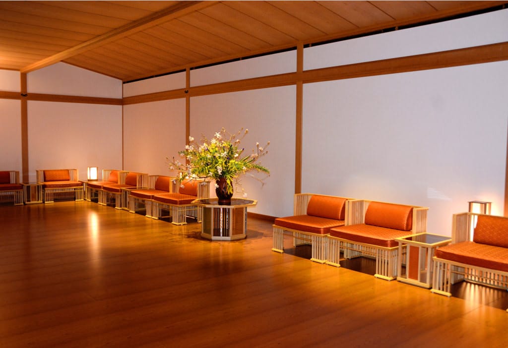 Une photo d’ensemble de la salle Juraku no Ma prise en biais. Des chaises rouge vif sont alignées contre les murs. Sur le piédestal, au centre, des fleurs sont disposées dans un panier à fleurs. 