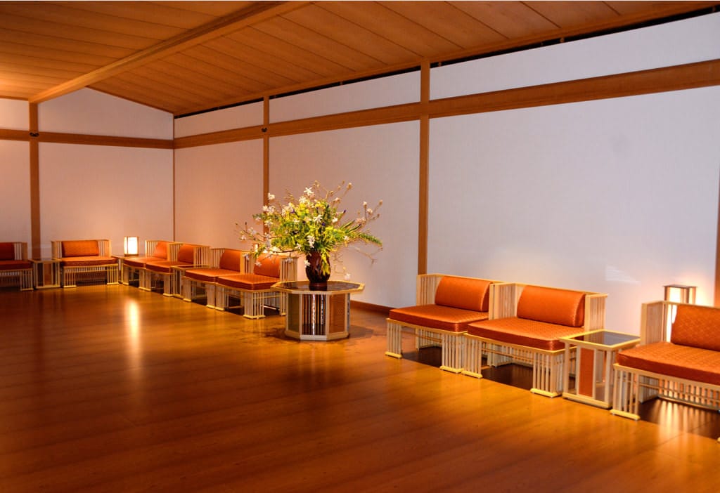 A photo of the whole of Juraku no Ma, photographing the room diagonally. Bright red chairs are lined up against the walls. On the pedestal in the center flowers are arranged in a flower vase. 