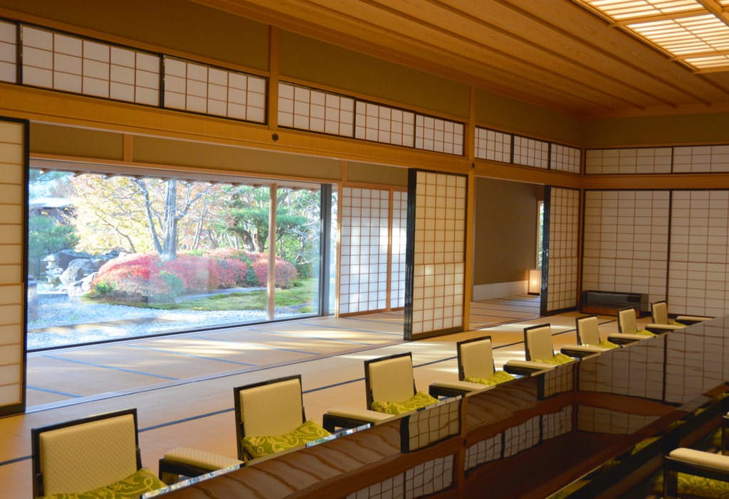 La salle de banquet de style japonais, « Kiri no Ma », photographiée en biais. Le sol est recouvert de tatamis et des chaises laquées sans pieds sont disposées autour d'une table basse au centre. Au-delà des portes coulissantes en papier, on peut apercevoir le jardin à l'extérieur.