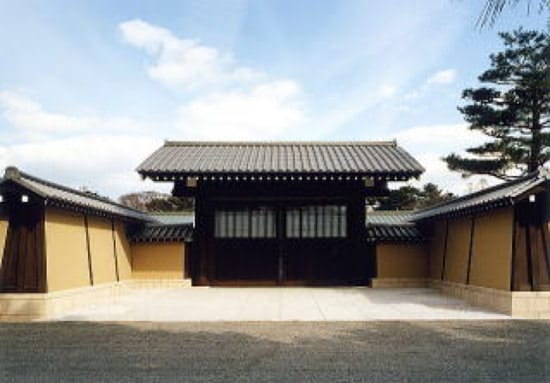 晴れた空を背景に、左右を築地塀に囲まれた京都迎賓館の正門が正面にあります。手前には京都御苑の砂利道があります。正門や、築地塀の後ろには木々が見えます。