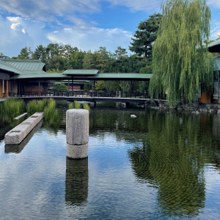 A photo of the Kyoto State Guest House's central garden, with a large pond in the center, willow tree to the right, and a covered bridge in the distance.