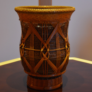 A woven flower vase is in the center of the photo. The vase's design is made with countless bamboo strips woven together. The vase is in the shape of a jar, but the top is made to open wide to accomodate flowers.