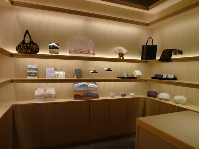 柔らかく照らされた3段の木製棚に、バッグ、クリアーファイル、扇子など京都迎賓館記念品が展示されている写真。