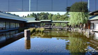 A photo of the garden, as viewed from the corridor in Yubae no Ma. Rattan blinds are hung across the top of the photo. Straight ahead, across the garden, a bridge crosses between the eastern and western eaves against a background of blue skies. The pond is fenced in by the building around it, and blue skies are reflected within it. Trees grow lush and green here.