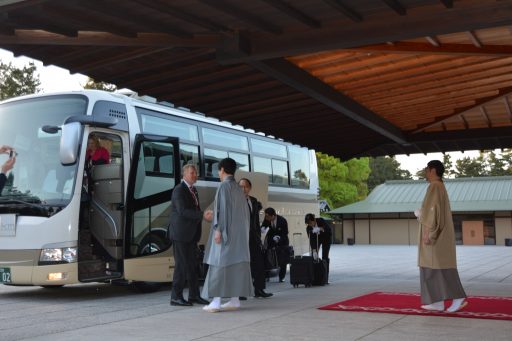 The delegation is getting off the bus and greeted by the Vice Director of the State Guest House with a handshake at the Main Entrance.